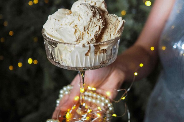 Woman in sparkly dress holding ice cream in a glass container wrapped with fairy lights and pearls, with a decorated Christmas tree lit with fairy lights in the background.